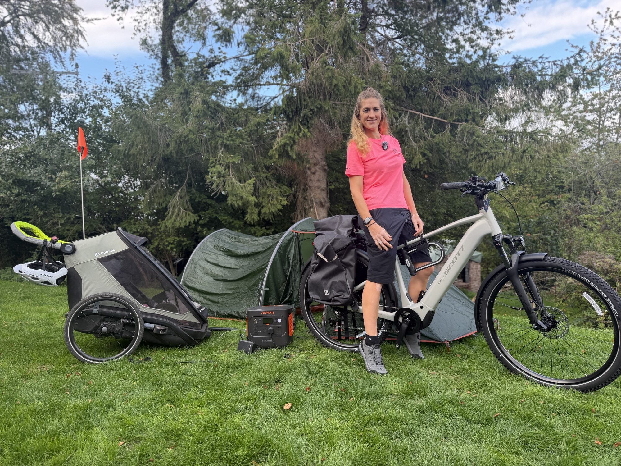 A cyclist standing beside a loaded SCOTT bike, child trailer and camping tent on grass.