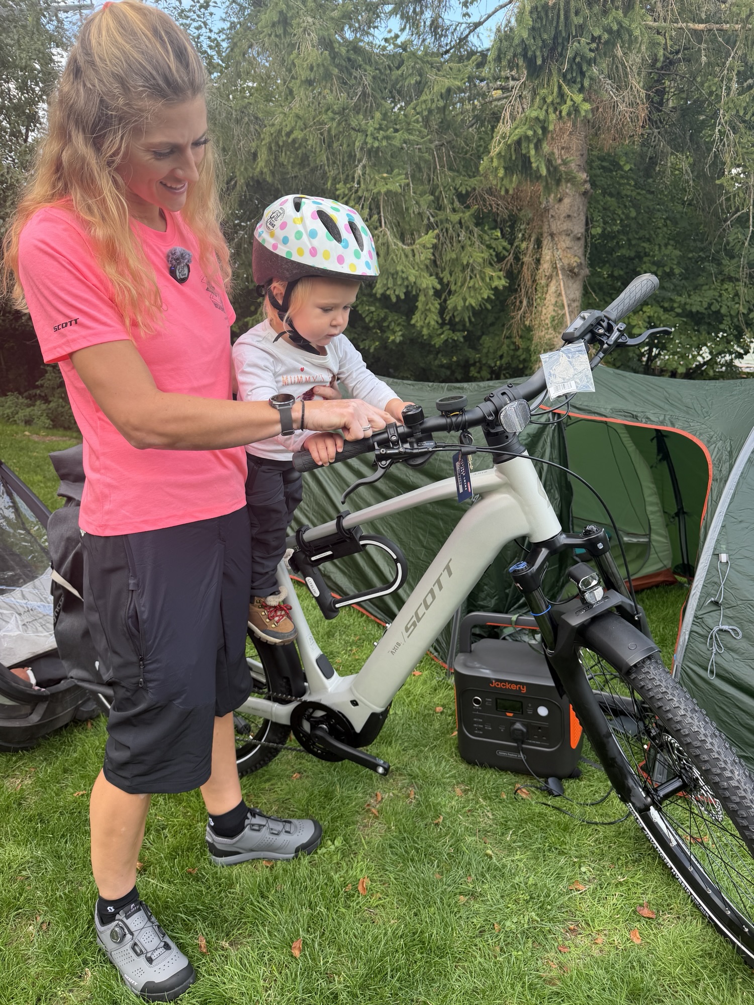 A toddler wearing a cycling helmet seated in front of an adult on a white SCOTT bike.