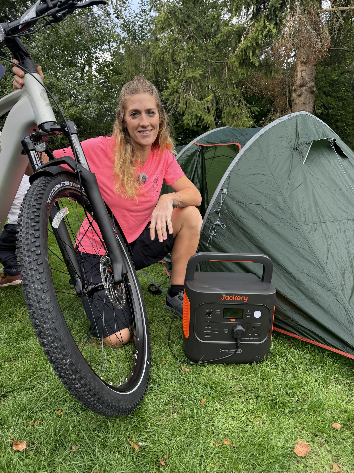 A cyclist kneeling beside a Jackery portable power station, SCOTT bike and green tent.