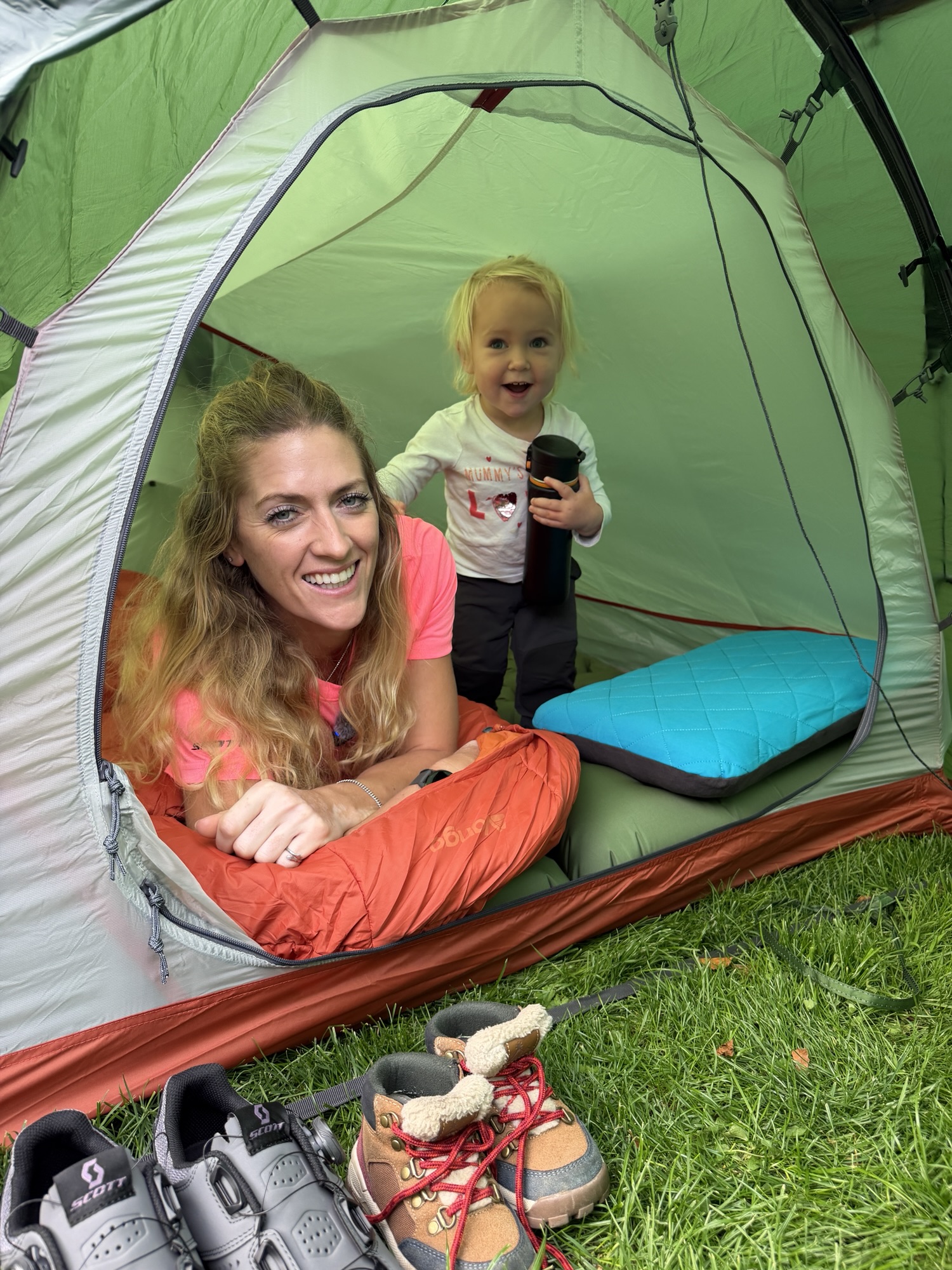 An adult lying in an orange sleeping bag inside a green tent with a toddler and a blue sleeping mat.